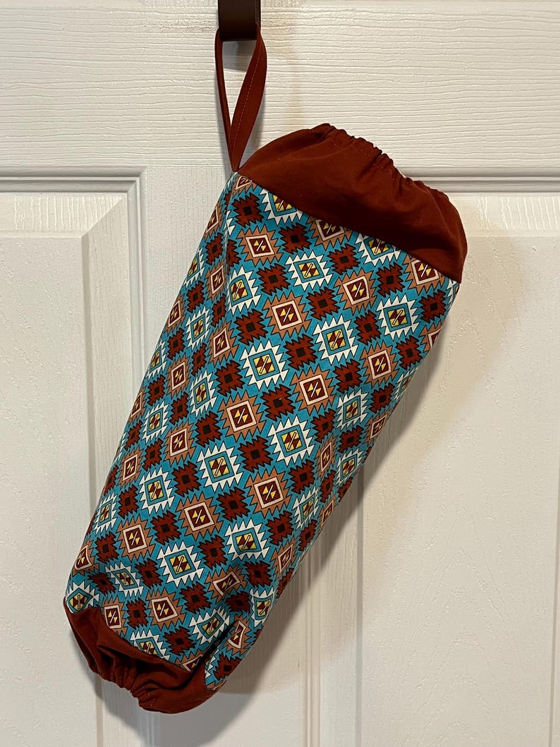 May include: A cylindrical fabric bag with a geometric pattern in teal, brown, and white. The bag has a drawstring closure and a loop for hanging. The bag is hanging on a brown hook.