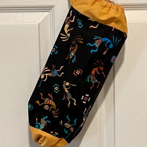 May include: A black fabric bag with a drawstring closure and a loop for hanging. The bag is patterned with Kokopelli figures in various colors. The top and bottom of the bag are a solid mustard yellow color. The bag is hanging on a white door.