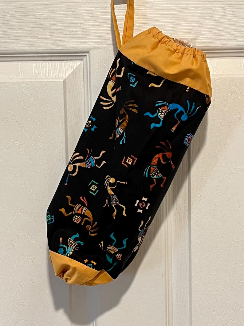 May include: A black fabric bag with a drawstring closure and a loop for hanging. The bag is patterned with Kokopelli figures in various colors. The top and bottom of the bag are a solid mustard yellow color. The bag is hanging on a white door.