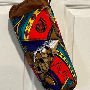 May include: A cylindrical fabric bag with a brown drawstring top and handle. The bag's body features a colorful pattern with Native American-inspired motifs in red, blue, yellow, and brown. The bag is hanging against a white door.