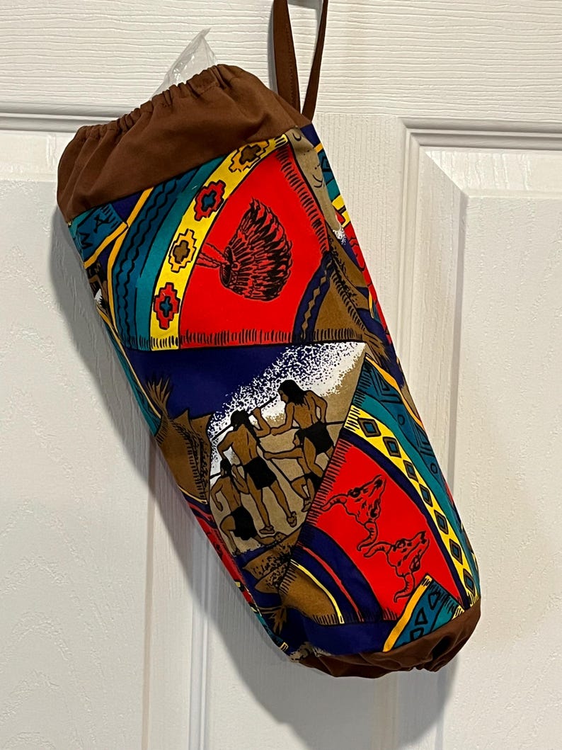 May include: A cylindrical fabric bag with a brown drawstring top and handle. The bag's body features a colorful pattern with Native American-inspired motifs in red, blue, yellow, and brown. The bag is hanging against a white door.