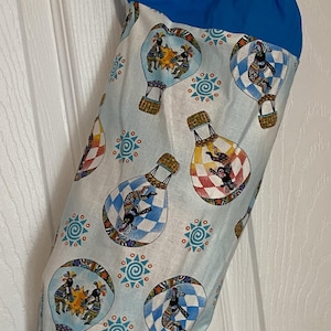May include: A blue and white fabric bag with a drawstring closure. The bag features a repeating pattern of hot air balloons with figures and geometric designs. The top and bottom of the bag are solid blue, and it has a blue loop for hanging.