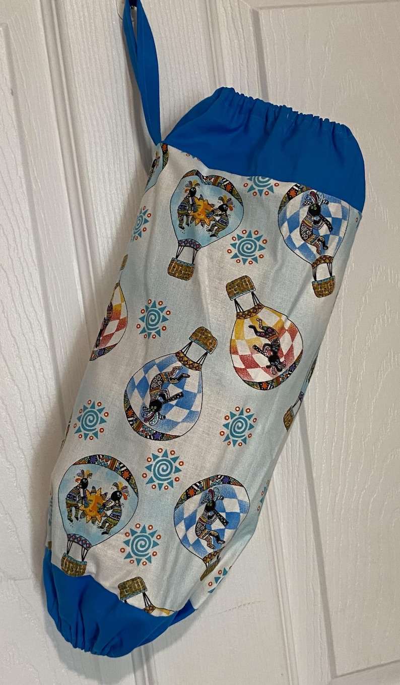 May include: A blue and white fabric bag with a drawstring closure. The bag features a repeating pattern of hot air balloons with figures and geometric designs. The top and bottom of the bag are solid blue, and it has a blue loop for hanging.