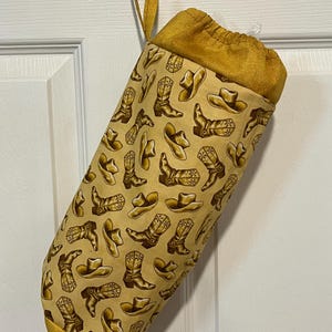 May include: A fabric stocking with a western theme. The stocking is a light tan color with a repeating pattern of cowboy boots and hats in shades of brown. The top and hanging loop are a solid gold color.