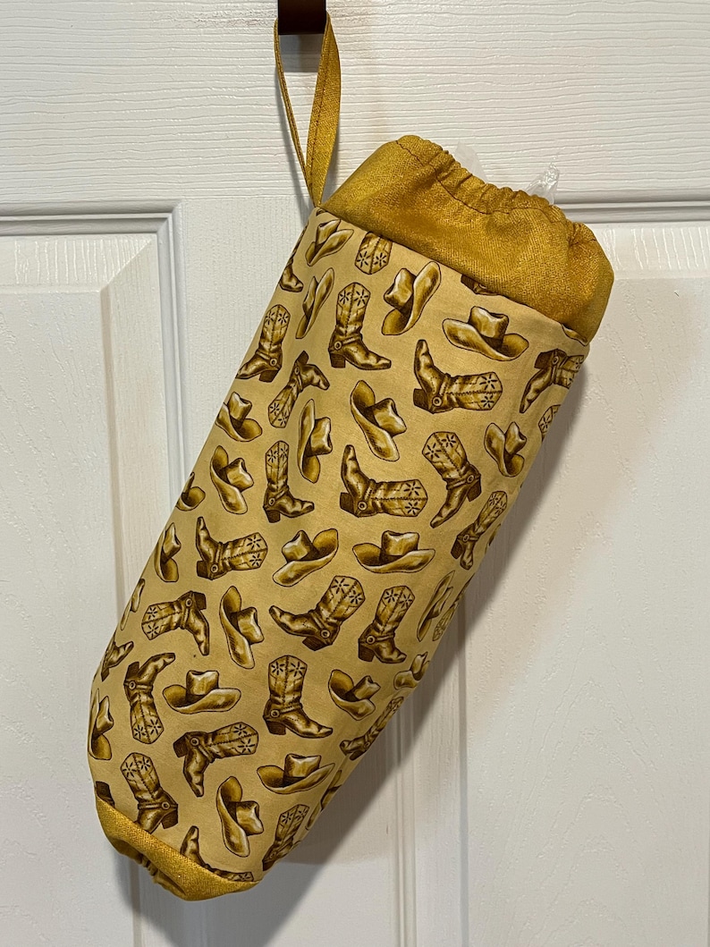 May include: A fabric stocking with a western theme. The stocking is a light tan color with a repeating pattern of cowboy boots and hats in shades of brown. The top and hanging loop are a solid gold color.