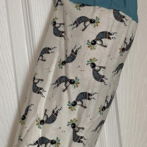 May include: A fabric bag with a teal top and bottom, and a white body. The white body is patterned with black, blue, green, and red Kokopelli figures. The bag is hanging from a hook.