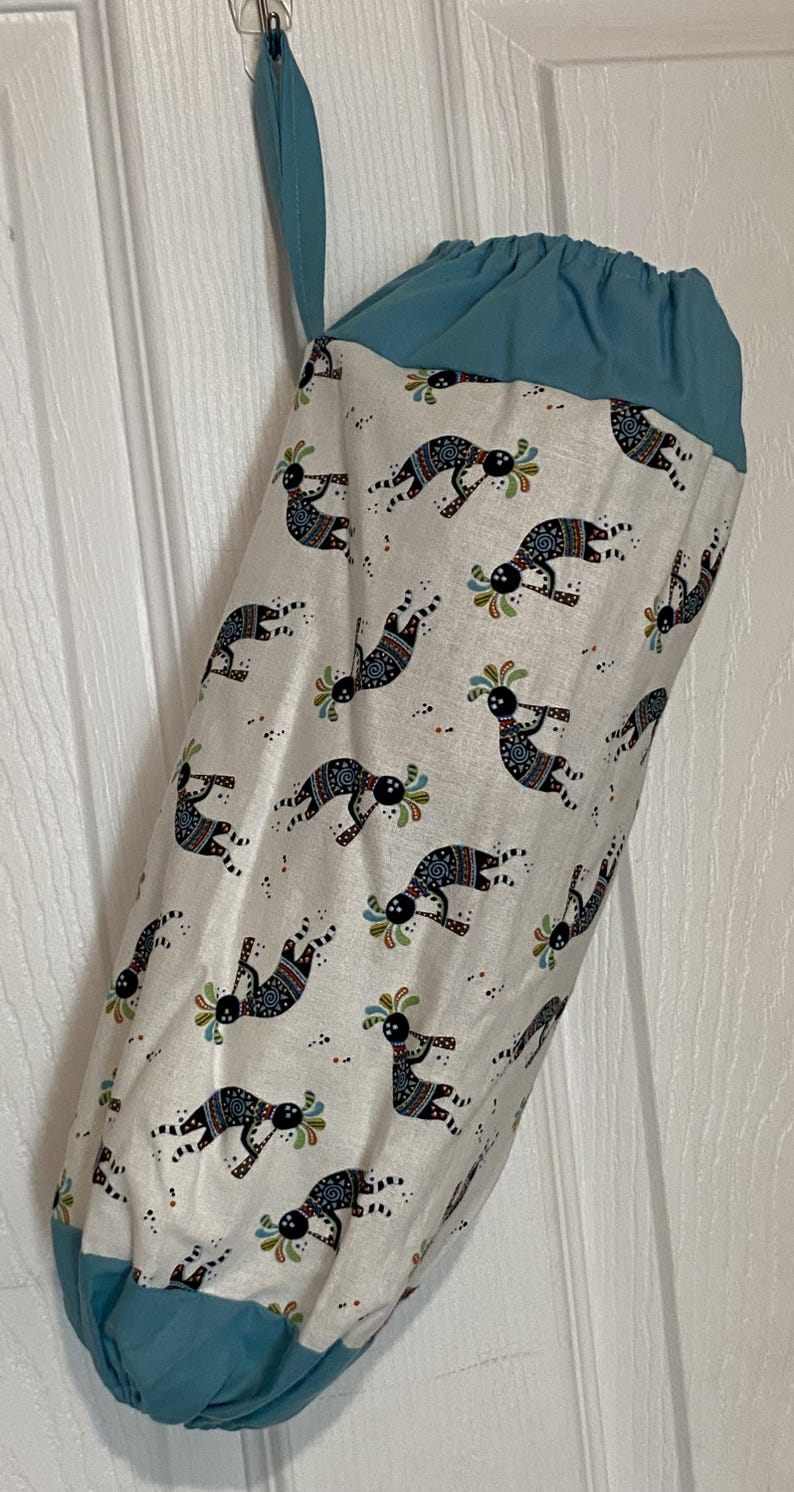 May include: A fabric bag with a teal top and bottom, and a white body. The white body is patterned with black, blue, green, and red Kokopelli figures. The bag is hanging from a hook.