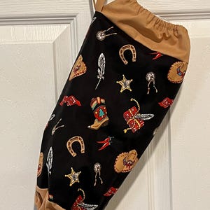 May include: A black fabric bag with a tan drawstring top and handle. The bag is patterned with cowboy boots, horseshoes, stars, feathers, and other Western-themed designs in red, gold, and white.