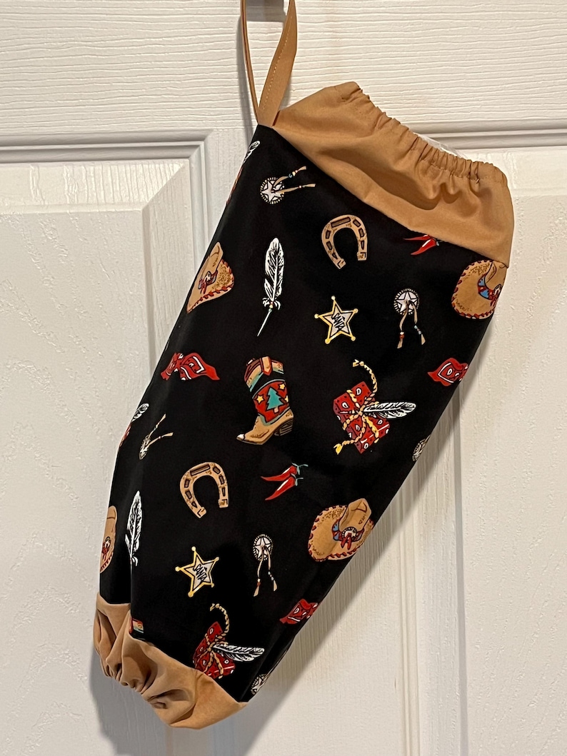May include: A black fabric bag with a tan drawstring top and handle. The bag is patterned with cowboy boots, horseshoes, stars, feathers, and other Western-themed designs in red, gold, and white.