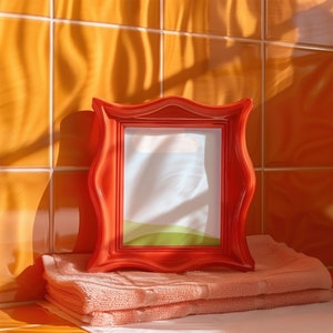 May include: A red picture frame with a wavy design sits on a stack of pink towels. The frame has a white mat and a clear window with a light blue sky and green grass.