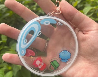 May include: A clear, round keychain with a rose gold clasp. The keychain features a blue and white design at the top and several colorful cartoon images inside, including a red creature, a blue jellyfish, and a green bucket.