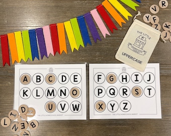 May include: Two white cards with black circles containing the uppercase alphabet letters A through Z. The cards are on a wooden table with a rainbow felt garland and a small white bag with the text "The Little Homeschool Market Uppercase" printed on it. There are wooden letter tiles scattered around the cards.