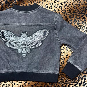 May include: A dark gray denim jacket with a black collar, cuffs, and hem. A large, detailed moth patch is centered on the back. The moth is primarily white with black and gray accents. The jacket has a bomber style.