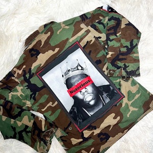 May include: A camouflage green jacket with a black and white image of a man wearing a crown with the word "Notorious" in red across his face.