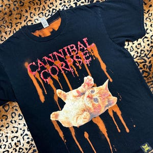 May include: Black t-shirt featuring the text "CANNIBAL CORPSE" in pink and orange dripping paint. A cartoon orange tabby cat is printed below the text. The shirt has a small yellow logo on the bottom right.