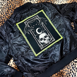 May include: A black bomber jacket featuring a rectangular patch on the back. The patch displays a white graphic of a skull, a crescent moon, and a winged creature, all framed by a border with bright yellow stitching on a black fabric background.