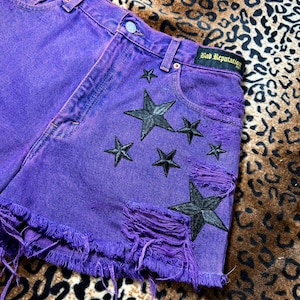 Può includere: Pantaloncini di jeans viola con dettagli strappati e toppe a forma di stella nera. I pantaloncini hanno un orlo sfilacciato e il testo "Bad Reputation" sulla tasca posteriore.