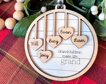 May include: A round wooden ornament featuring a white shiplap-style background with four hanging heart-shaped cutouts engraved with the names Will, Joey, Lucy, Evan, and Liam. The text Grandchildren make life grand is printed in black.