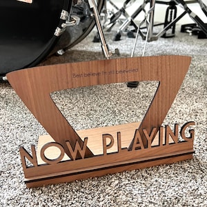 May include: A wooden sign with the words "NOW PLAYING" in large letters. The sign is made of dark wood and has a black border. The sign is sitting on a brown carpet. The text "Best believe I'm still bejeweled" is engraved on the top of the sign.