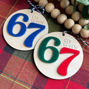 May include: Two circular wooden ornaments featuring raised acrylic numbers. One ornament displays a blue number 67 with the text SIX SEVEN. The other ornament displays a green number 6 and a red number 7 with the text SIX SEVEN. Both have hanging strings.