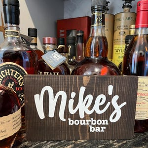 May include: A dark stained wooden sign with the words "Mike's bourbon bar" in white lettering. The sign is rectangular and stands in front of several bottles of bourbon.