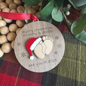 May include: Wooden Christmas ornament with the text "merry christmas mommy & daddy" and "see you in 2026". The ornament features a Santa hat, baby footprints, and snowflakes. A red ribbon is attached for hanging.