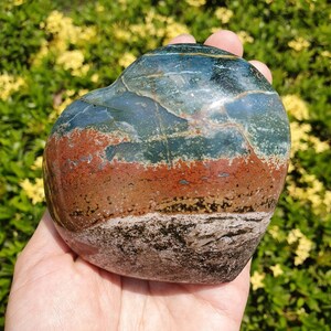 May include: A heart-shaped polished stone with bands of color. The stone features layers of dark teal, rust, brown, and white. The stone is held in a hand, with a blurred green and yellow background.