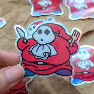 Może przedstawiać: Zestaw holograficznych naklejek przedstawiających postać z kreskówki w czerwono-białym stroju, trzymającą nóż i widelec. Postać ma białą twarz z czarnymi oczami i zaskoczony wyraz twarzy.