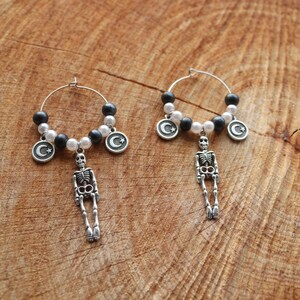 May include: Silver hoop earrings with a gothic design. Each earring has a skeleton charm, black and white beads, and a crescent moon and star charm. The earrings are presented on a wooden surface, showcasing the intricate details.