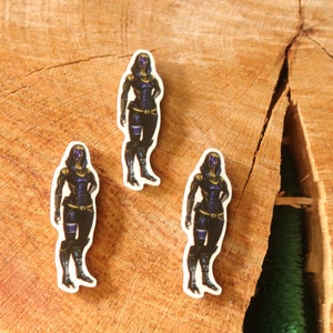 May include: Three stickers featuring a character in a black and blue outfit with a helmet. The stickers are placed on a wooden surface with a natural wood grain pattern. The character has a futuristic design.