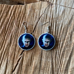 May include: A pair of round earrings featuring a portrait of a character with a pale face and pins. The earrings have a white border and silver hooks. The background is a dark blue gradient.