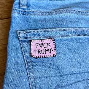 May include: Close-up of light blue denim jeans with a back pocket. A pink patch with black text that reads "FVCK TRUMP" is sewn onto the pocket. The patch has a stitched border.
