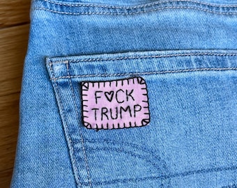 Può includere: Primo piano di jeans blu chiaro con una tasca posteriore. Una toppa rosa con testo nero che recita "FVCK TRUMP" è cucita sulla tasca. La toppa ha un bordo cucito.