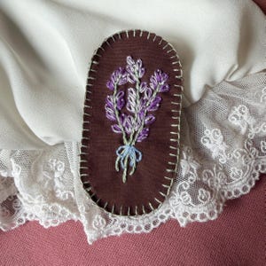 May include: A brown, oval-shaped embroidered patch featuring a bouquet of lavender. The lavender is purple and green, tied with a light blue ribbon. The patch is stitched with white thread around the edges.