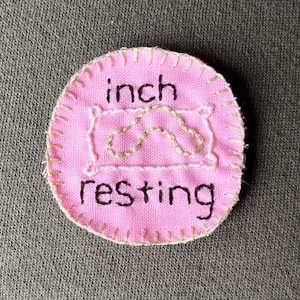 May include: A pink fabric patch with the words "inch resting" embroidered in black thread. The patch features a white outline of a pillow with a beige embroidered shape inside.