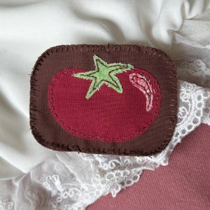 May include: A handmade, rectangular patch featuring a red tomato with a green stem and a brown border. The tomato is stitched onto a brown background. The patch is likely for clothing or accessories.