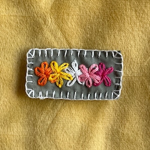 May include: A gray fabric patch with a white stitched border. Five embroidered flowers in a row, orange, yellow, white, pink, and red.