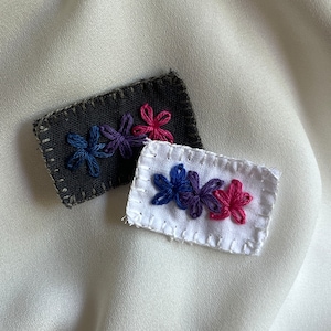 May include: Two embroidered fabric patches with flower designs. The first patch is dark gray with three purple and blue flowers. The second patch is white with three flowers, one pink, one purple, and one blue.