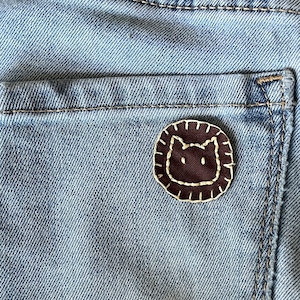May include: A brown cat face embroidered patch with white stitching on a blue denim background.
