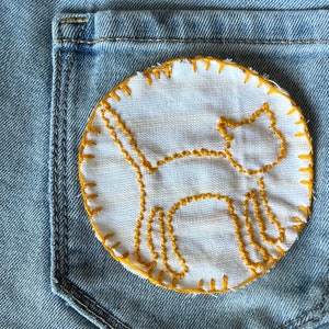 May include: A white fabric patch with a yellow embroidered cat design sewn onto a blue denim pocket.