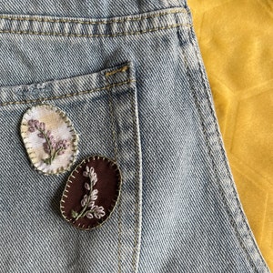 May include: Two embroidered patches sewn onto a light blue denim fabric. The patches are oval shaped and feature a floral design in shades of pink and white.
