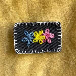 May include: A black fabric patch with a white stitched border featuring three embroidered flowers in blue, yellow, and pink.