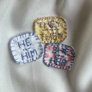 May include: Three square fabric patches with embroidered text. The patches are sewn with blue thread. The text on the patches reads "He/Him", "She/Her", and "They/Them".