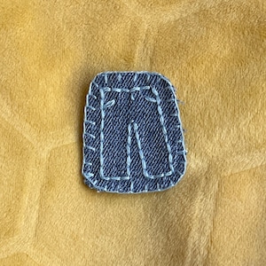 May include: A small, blue denim patch in the shape of a pair of pants, with white stitching around the edges.