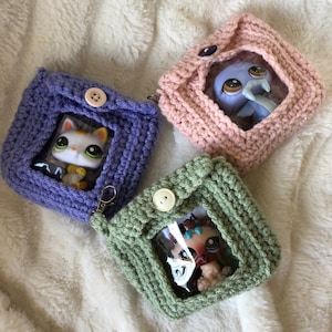 Puede incluir: Tres bolsas de ganchillo con ventanas de plástico transparente, cada una con un juguete pequeño diferente. Las bolsas son moradas, rosas y verdes.