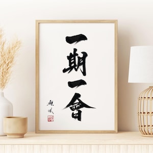 Puede incluir: Obra de arte enmarcada con caligrafía japonesa en tinta negra sobre fondo blanco. La caligrafía dice "Ichi-go ichi-e", que significa "una vez, un encuentro". La obra de arte está en un marco de madera clara, exhibida en un estante blanco.