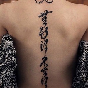 Puede incluir: Un tatuaje de tinta negra de una l&iacute;nea vertical de caligraf&iacute;a en la espalda de una persona. El tatuaje est&aacute; en la parte baja de la espalda y se extiende hasta el cuello. Hay un tatuaje m&aacute;s peque&ntilde;o de un s&iacute;mbolo de infinito con n&uacute;meros en el cuello.