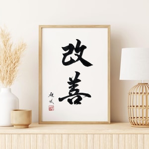 K&ouml;nnte beinhalten: Gerahmter Druck mit schwarzer Kalligraphie auf wei&szlig;em Hintergrund. Das Kunstwerk zeigt zwei gro&szlig;e chinesische Schriftzeichen. Links eine wei&szlig;e Vase mit getrocknetem Pampasgras und eine kleine beige Tasse. Rechts eine Lampe mit gewebtem Sockel.