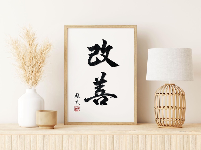 K&ouml;nnte beinhalten: Gerahmter Druck mit schwarzer Kalligraphie auf wei&szlig;em Hintergrund. Das Kunstwerk zeigt zwei gro&szlig;e chinesische Schriftzeichen. Links eine wei&szlig;e Vase mit getrocknetem Pampasgras und eine kleine beige Tasse. Rechts eine Lampe mit gewebtem Sockel.