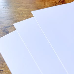 K&ouml;nnte beinhalten: Drei Bl&auml;tter wei&szlig;es Papier sind aufgef&auml;chert, ihre Kanten sind auf einer Holzoberfl&auml;che sichtbar. Das Papier ist strahlend wei&szlig; und das Holz hat einen warmen, braunen Ton. Die Komposition deutet auf Schreibwaren oder Kunstbedarf hin.